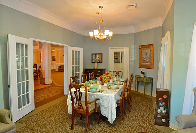 A formal dining room with a rectangular table covered with a white lace tablecloth set for eight people with plates, cups, and utensils. The room has light blue walls, a chandelier hanging from the ceiling, and French doors leading to another room. There is a small table with a flower vase and a framed painting on the wall.