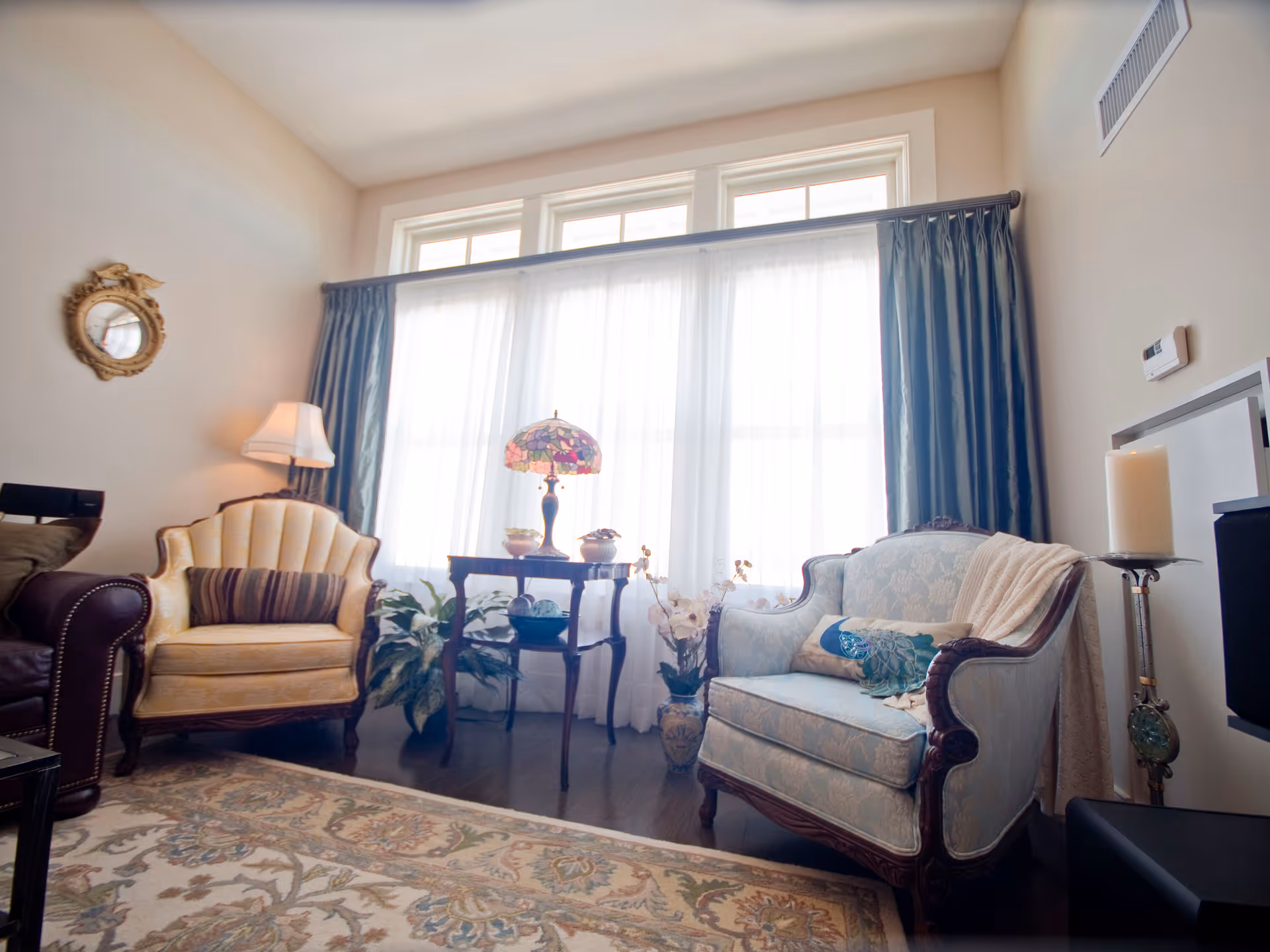A cozy living room with two vintage armchairs, one yellow and one light blue, separated by a small wooden table with a decorative lamp and plants. The room has large windows with sheer white curtains and dark blue drapes, a patterned area rug, a wall-mounted thermostat, and a candle on a tall stand.