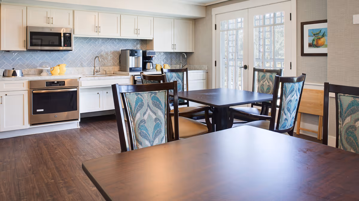 A bright kitchen and dining area with white cabinets, stainless steel microwave and oven, coffee machines on the counter, and wooden tables with chairs that have patterned upholstery. French doors with glass panes allow natural light into the room.