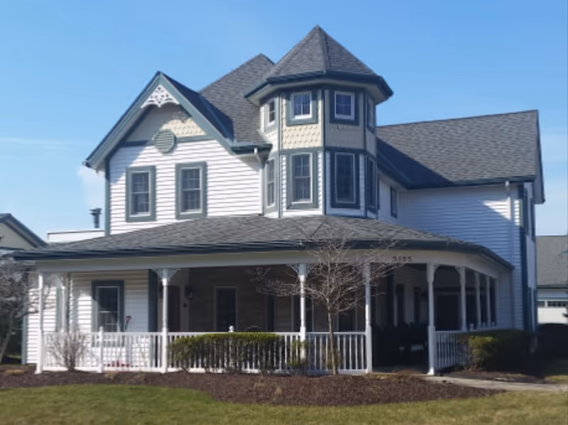A large, two-story white house with gray trim and a wraparound porch. The house features a turret with multiple windows and a steeply pitched roof. There is a small tree and some bushes in front of the porch, and the lawn is green under a clear blue sky.