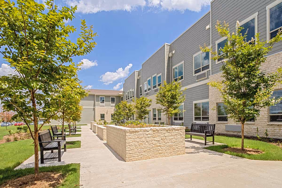 Outdoor courtyard area of Midtowne Assisted Living and Memory Care featuring a paved walkway, several black benches, young trees, and raised stone planters under a blue sky with some clouds.