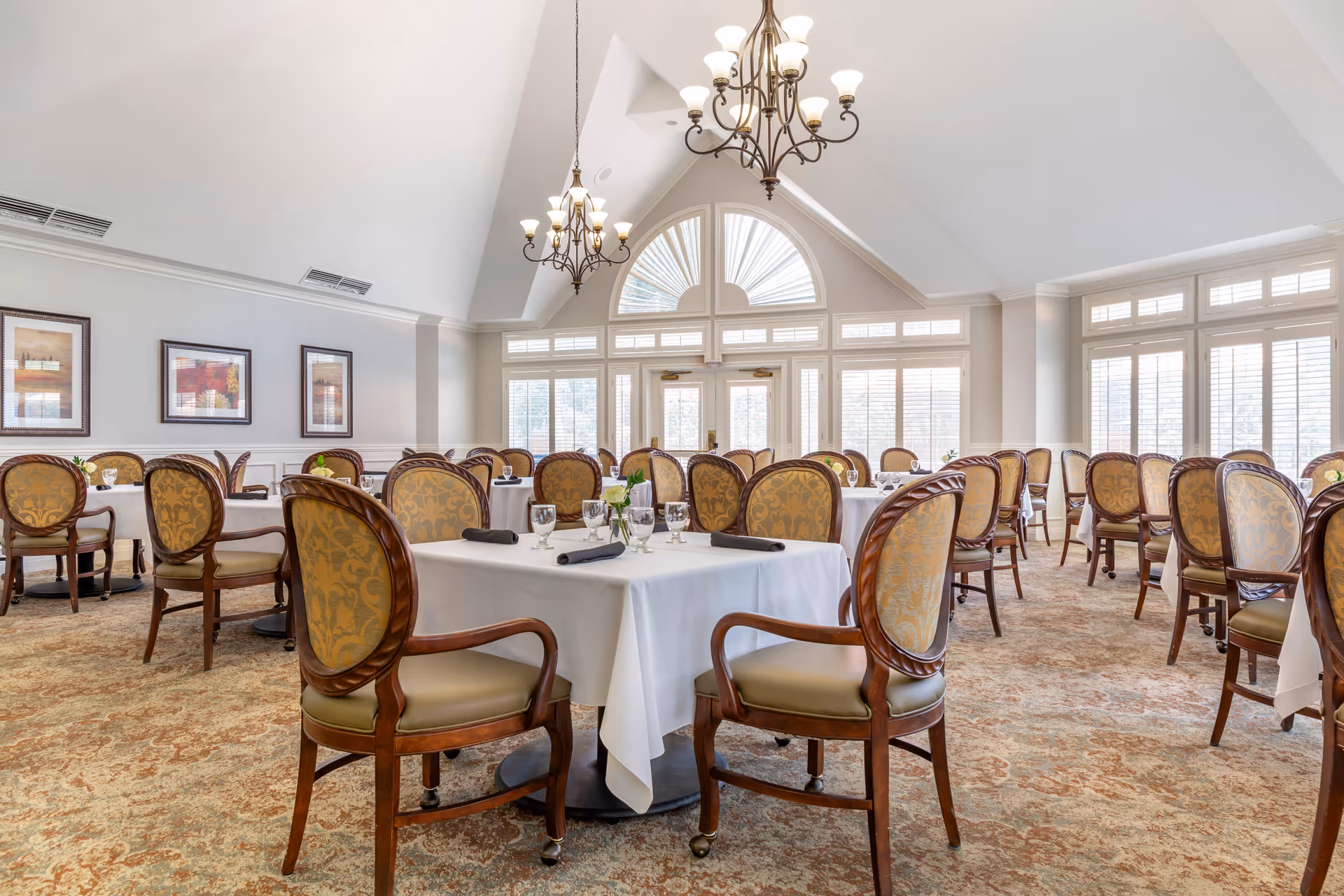 A bright, elegant dining room with round tables covered in white linens, upholstered wooden chairs, chandeliers, and large arched windows under a vaulted ceiling.