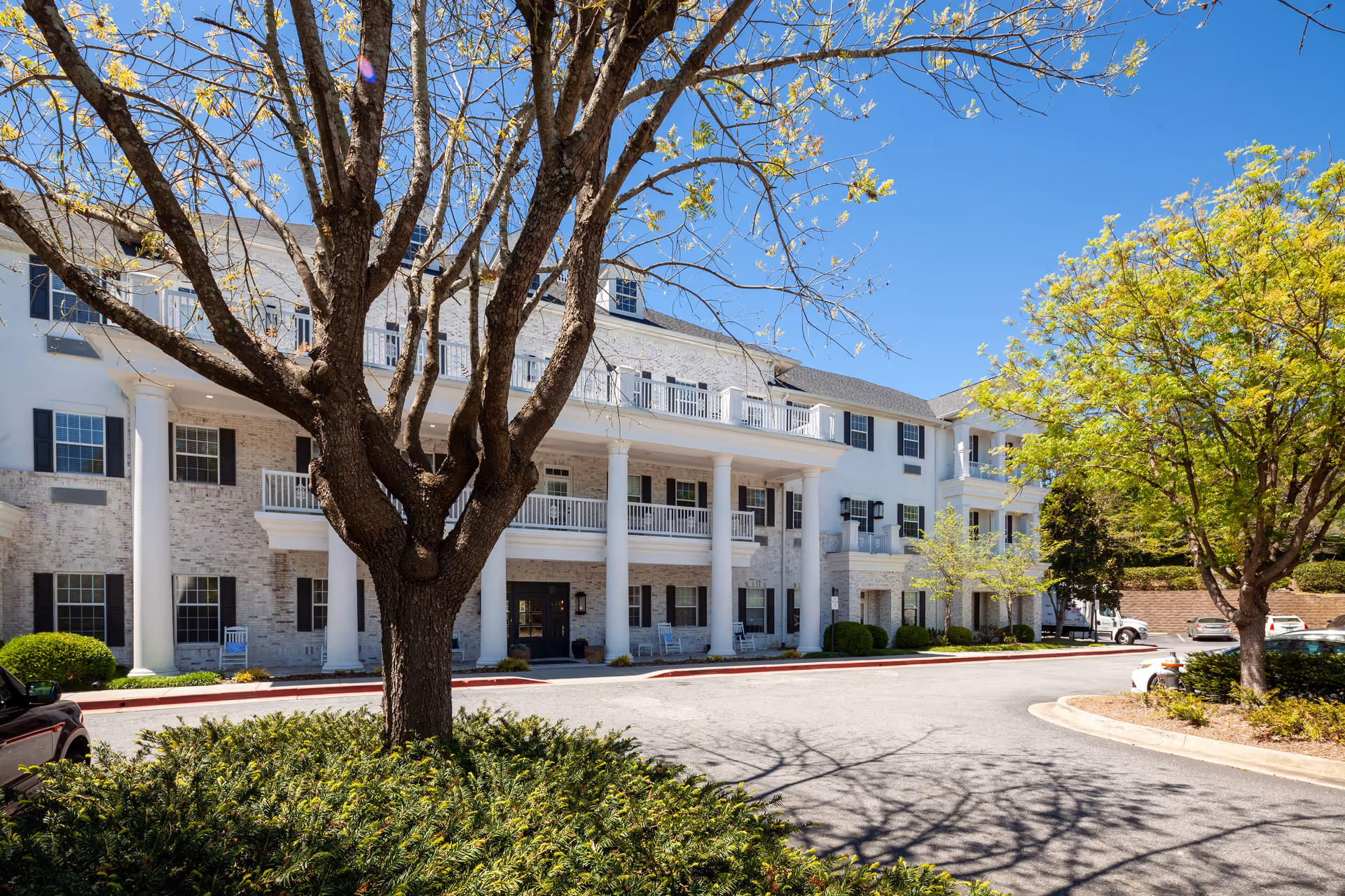 Exterior view of a large, white brick senior living facility building with multiple windows and balconies. The building has tall white columns supporting a covered entrance. There are trees with fresh green leaves and a clear blue sky in the background. A driveway and parked cars are visible in front of the building.