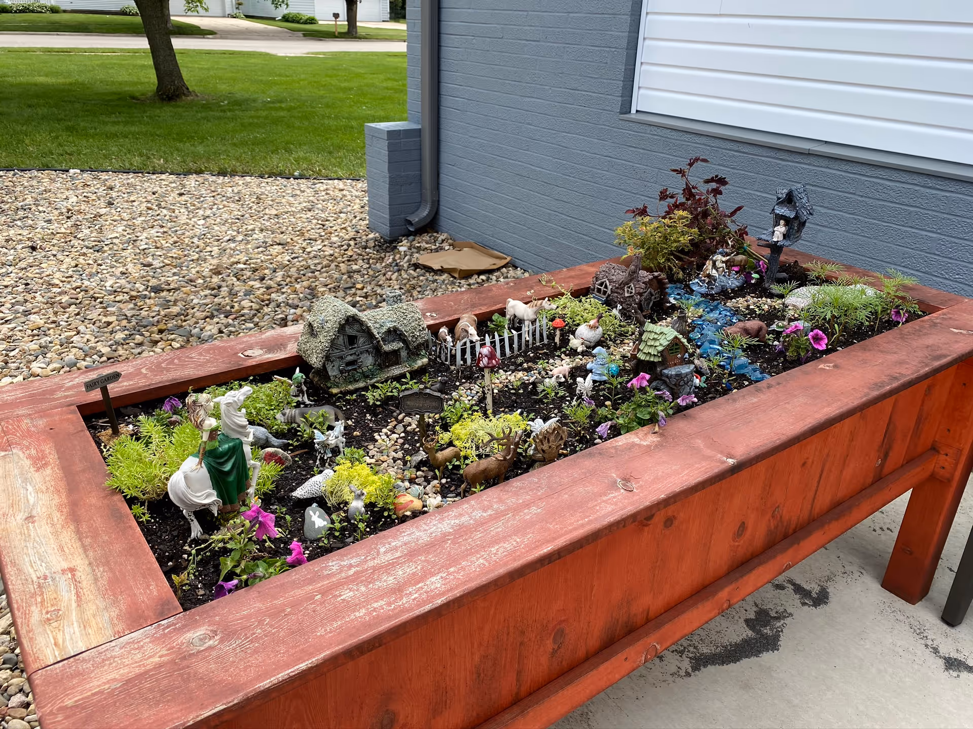 A raised wooden garden bed filled with soil, small plants, flowers, and various miniature decorative figurines including houses, animals, and a figure on a horse. The garden bed is placed outdoors on a concrete surface next to a building with gray and white siding. In the background, there is a lawn with green grass and trees.