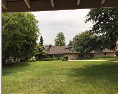 View of a single-story building with a brown roof surrounded by green trees and a well-maintained grassy lawn under an overcast sky, seen from under a shaded structure.
