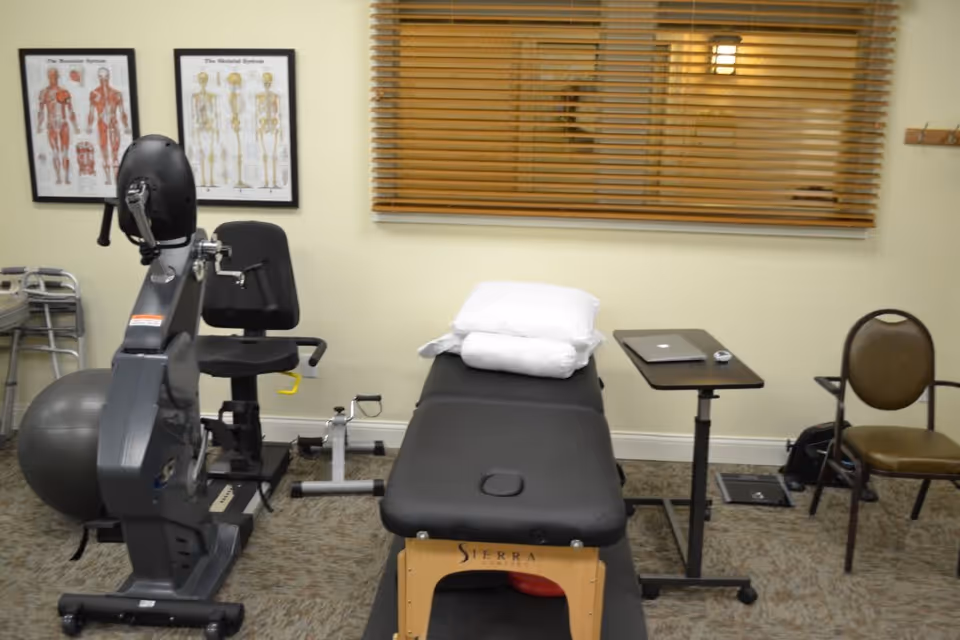 A therapy or rehabilitation room with exercise equipment including a stationary bike, a therapy table with folded white towels on top, a small adjustable table with a laptop, two anatomical posters on the wall, a window with wooden blinds, and a chair.