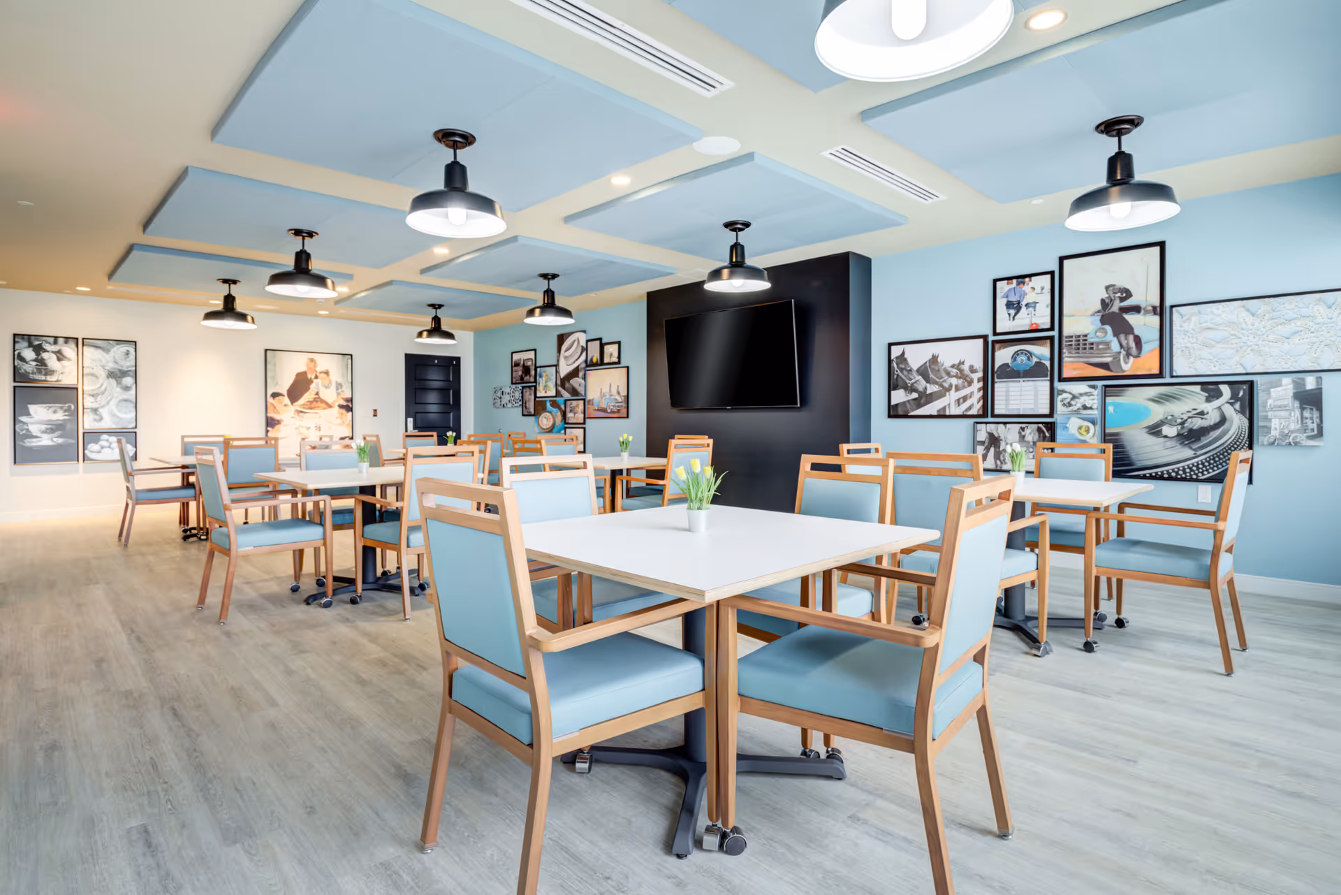 A bright and modern dining room with multiple square tables and wooden chairs with light blue cushions. The room features light wood flooring, light blue ceiling panels with black hanging lights, and a wall decorated with various framed pictures. A large flat-screen TV is mounted on a black accent wall.