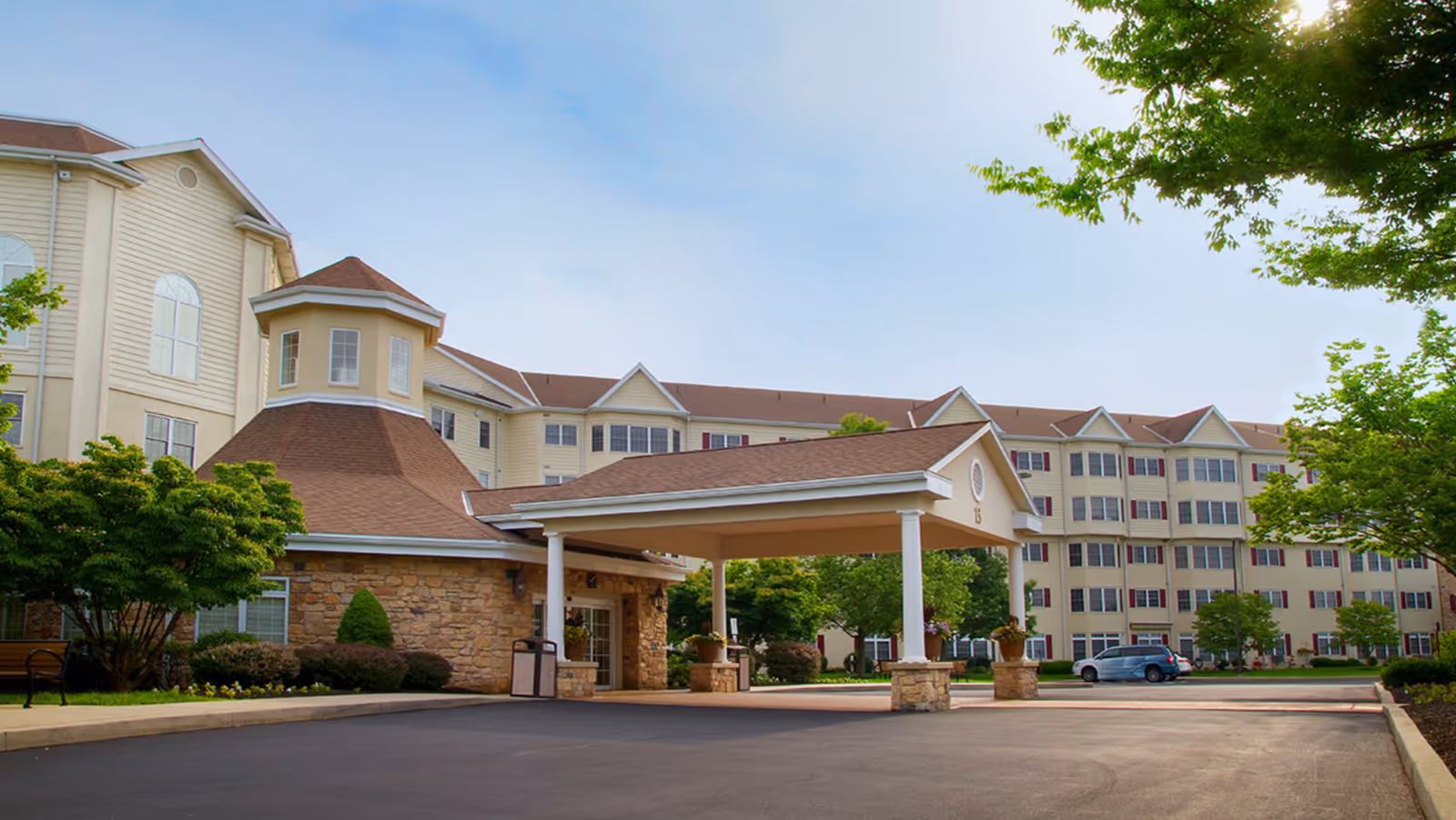 Exterior view of a large senior living facility building with beige siding and a brown roof. The entrance has a covered driveway supported by white columns and stone bases. There are trees and shrubs around the building and a clear blue sky above.