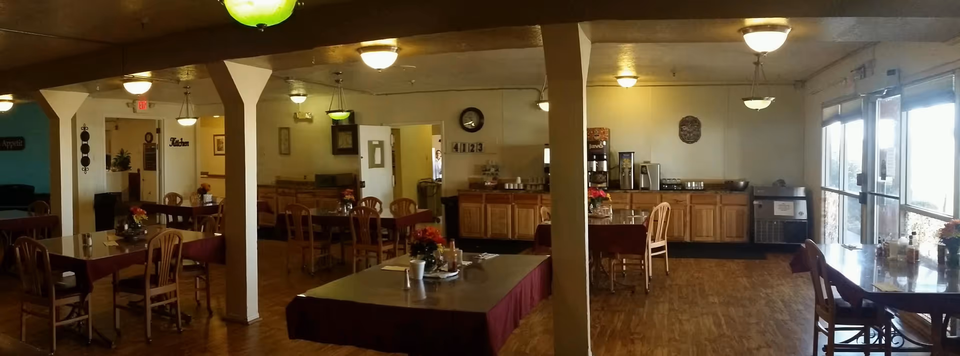 Interior view of a senior dining room with multiple wooden tables and chairs, a buffet counter along the back wall, and large windows on the right.