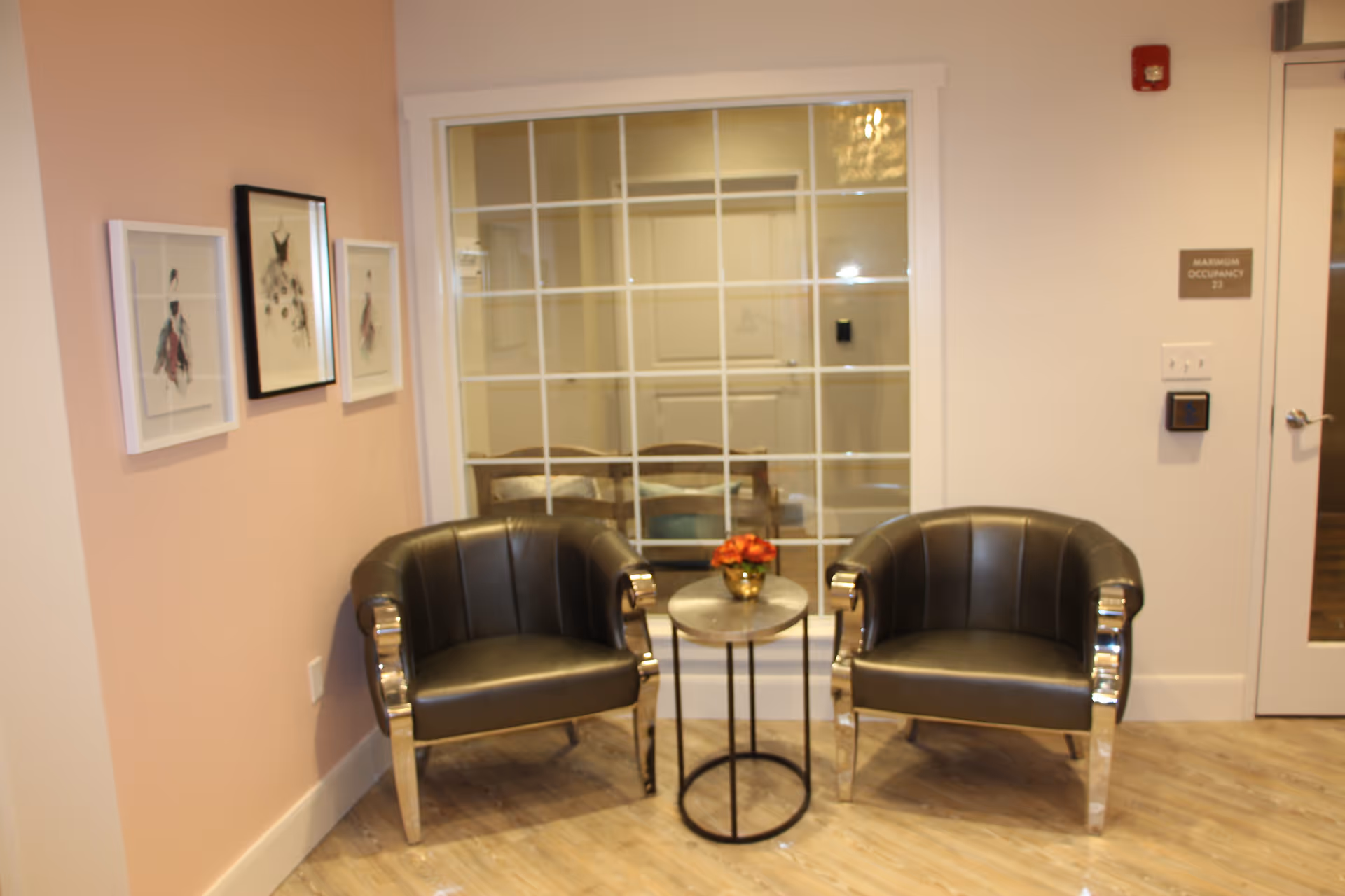 A cozy seating area with two black leather armchairs with shiny metal armrests and legs, separated by a small round table with a flower arrangement in a gold vase. The area has light-colored wooden flooring and a peach-colored wall with three framed fashion sketches. Behind the seating area is a large window with white grid framing, and to the right is a white door with a sign indicating maximum occupancy of 29.
