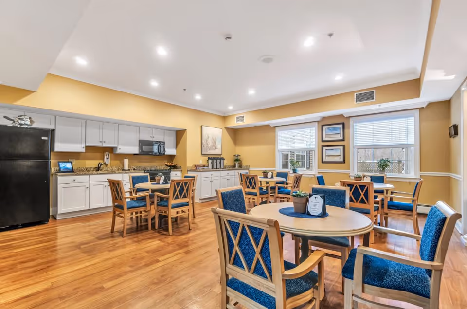 A bright dining room with wooden floors, several round tables surrounded by wooden chairs with blue cushions, and a kitchenette area with white cabinets, a black refrigerator, and a microwave. The walls are painted yellow, and there are windows with white blinds letting in natural light. Some framed artwork and small plants decorate the space.
