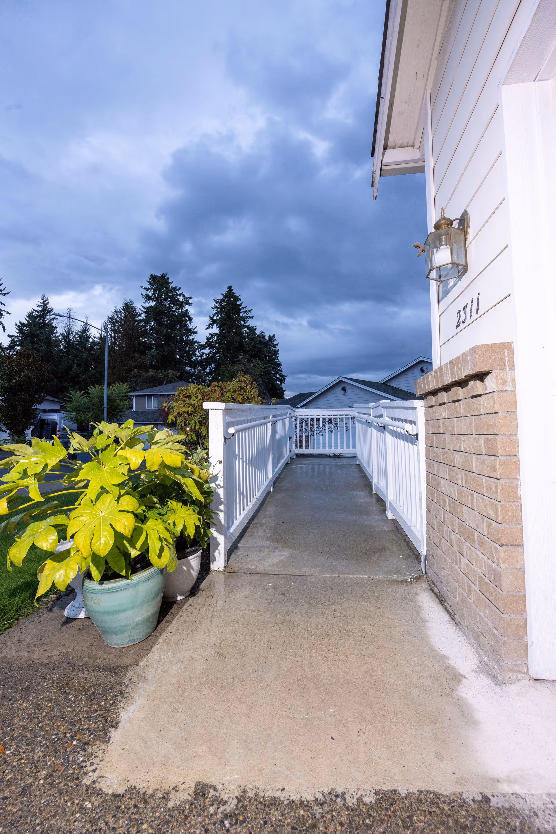 Outdoor view of a concrete ramp with white railings leading to the entrance of a building with the number 2511 on the wall. There are potted plants with green leaves on the left side of the ramp and trees and houses visible in the background under a cloudy sky.