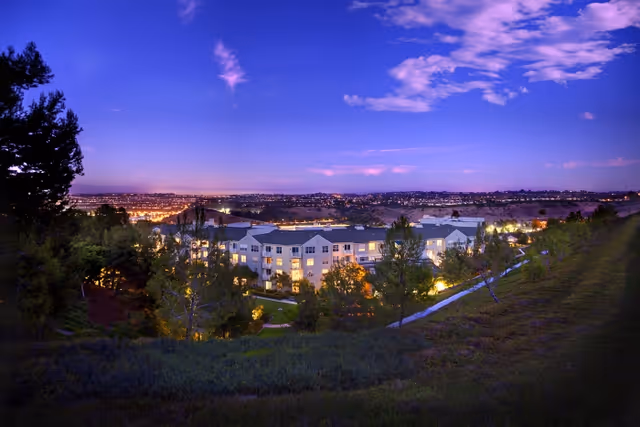 Evening view of The Covington senior living facility surrounded by trees and greenery, with city lights visible in the distance under a partly cloudy sky.