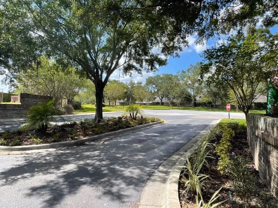 A sunny outdoor scene showing a curved driveway lined with landscaped flower beds and trees. There is a stop sign visible on the right side near the end of the driveway, and a brick wall on the right side. The sky is blue with some clouds, and shadows from the trees fall on the pavement.