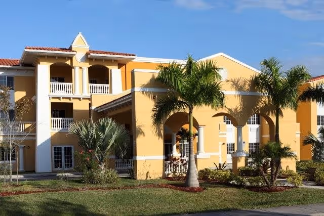 Exterior view of a yellow two-story assisted living community building with white trim, balconies, and arched entryways, surrounded by palm trees and landscaped greenery under a clear blue sky.