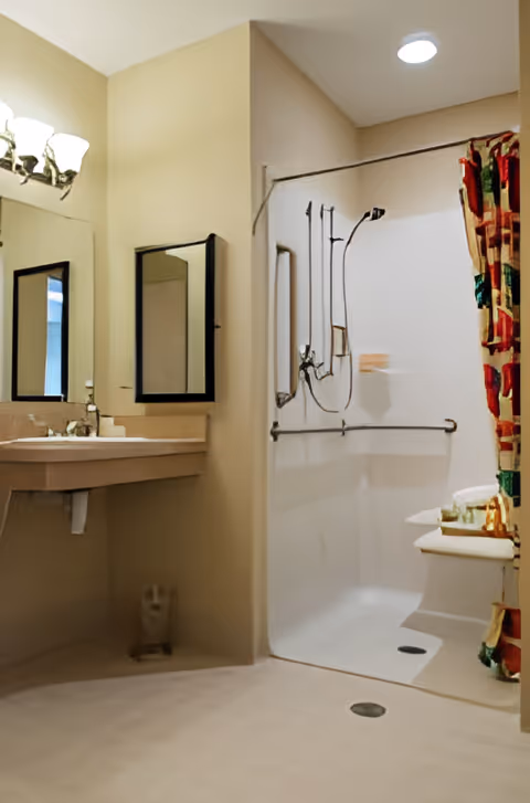 A bathroom with a beige color scheme featuring a wall-mounted sink with a mirror above it, a walk-in shower with grab bars, a handheld showerhead, and a colorful shower curtain. The floor is light-colored and there is a small drain visible.