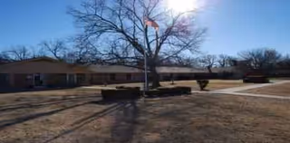 One-story beige nursing facility exterior with flagpole, trees, and benches in a courtyard.