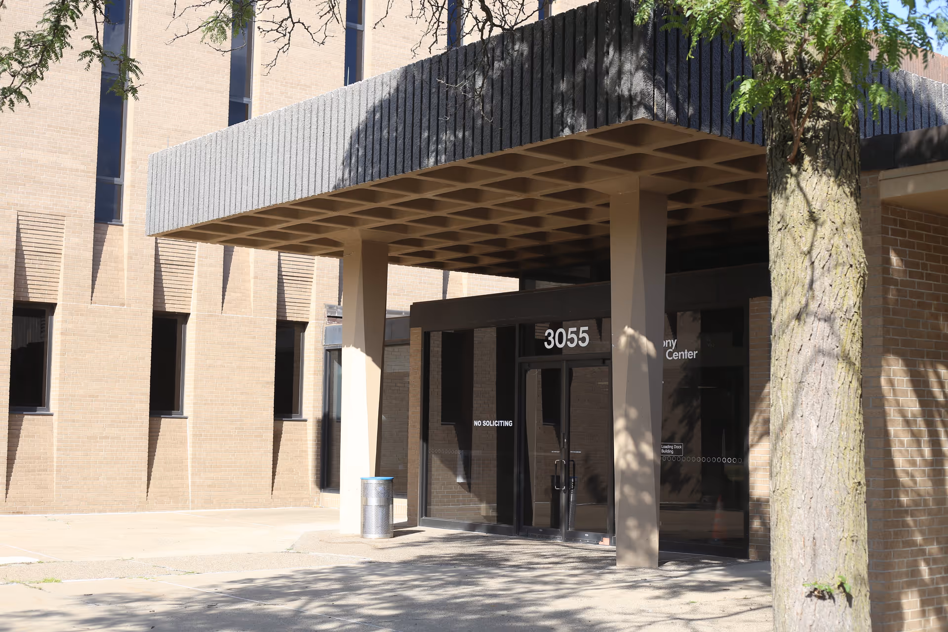Exterior view of a brick building entrance with the number 3055 above glass doors. The entrance has a large overhang supported by two columns, and there is a tree and a trash can nearby. The words 'NO SOLICITING' are visible on one of the glass doors.