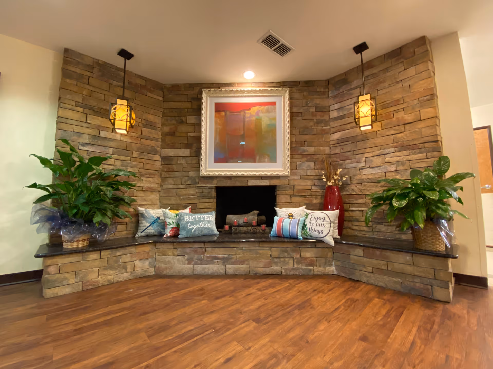 A cozy indoor seating area with a stone fireplace surrounded by a stone wall. The seating area has a black granite ledge with decorative pillows, including ones that say 'Better together' and 'Enjoy the little things.' There are two hanging pendant lights above, a large abstract painting above the fireplace, and two large green plants in baskets on either side.