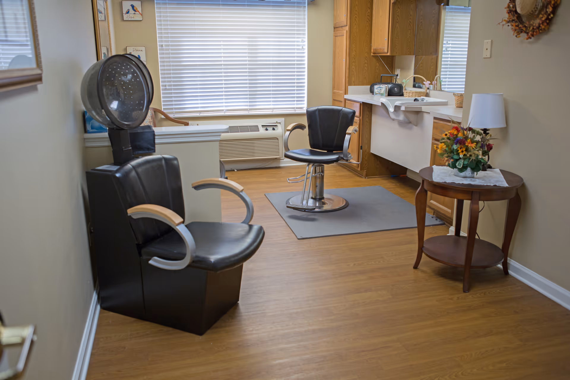 Small salon area with a hooded hair dryer chair and a styling chair by a sink and counter, with a side table holding a lamp and flowers.