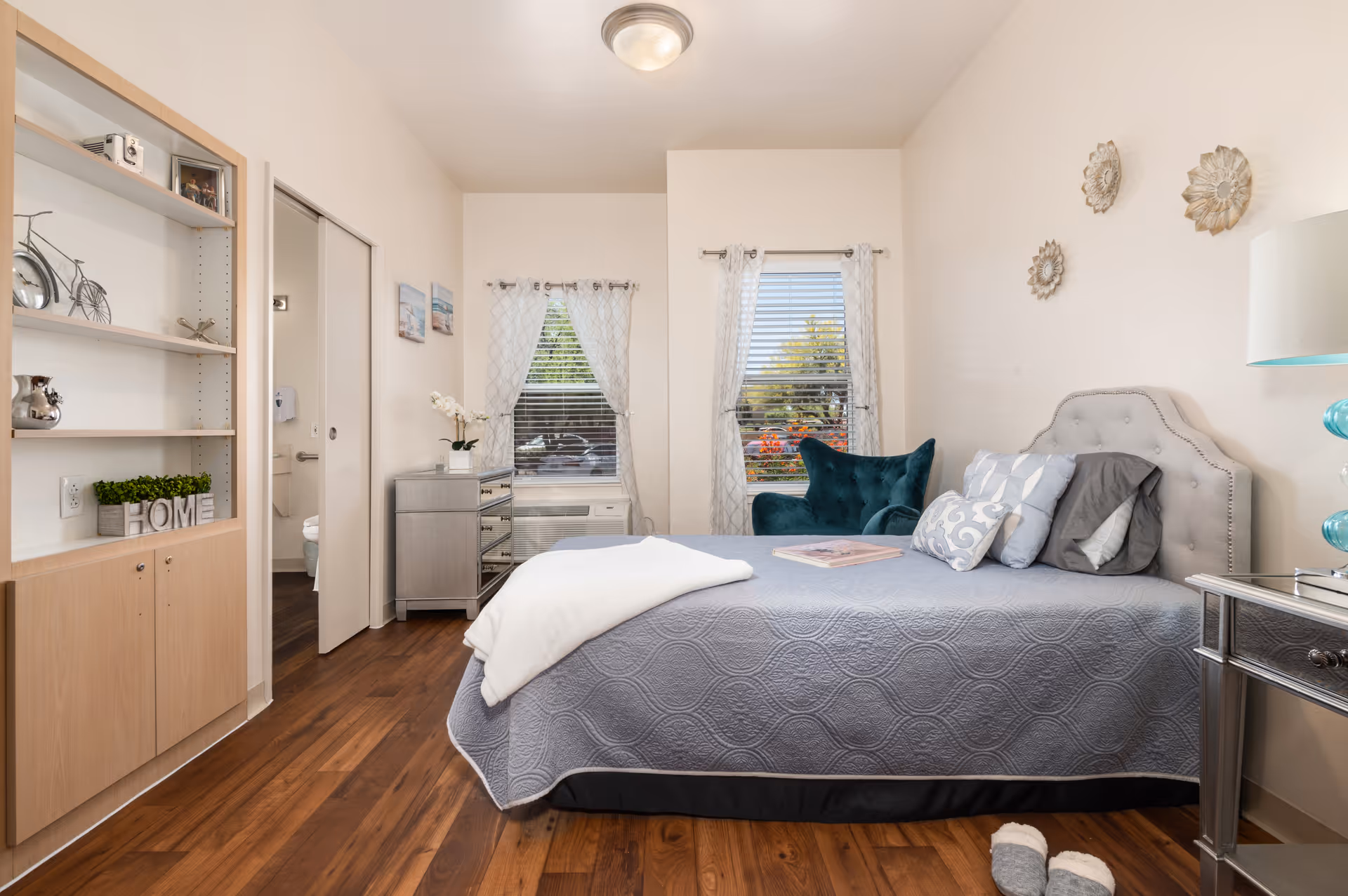 A cozy bedroom in an assisted living facility featuring a neatly made bed with gray bedding and multiple pillows, a teal armchair near two windows with white curtains, a mirrored nightstand with a lamp, a small dresser with a plant, and a built-in wooden shelving unit with decorative items. The room has wooden flooring and a door leading to a bathroom.