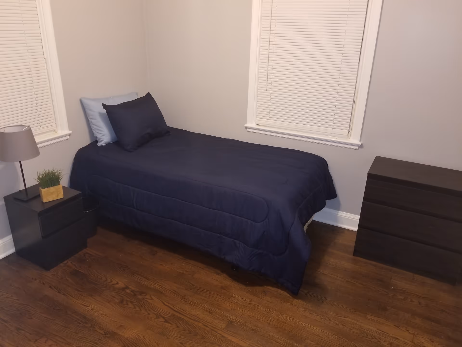 A small bedroom with a single bed covered in a dark blue comforter and two pillows, one light blue and one dark blue. There is a black nightstand with a lamp and a small plant on it next to the bed. Across from the bed is a black three-drawer dresser. The room has wooden floors and two windows with closed white blinds.