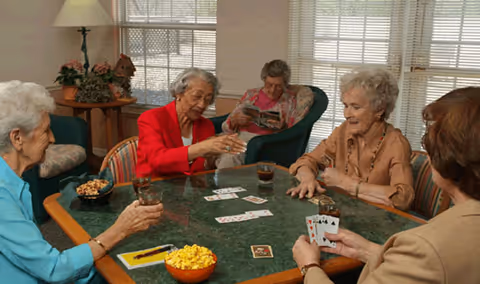 Four elderly women sitting around a green table playing cards and enjoying drinks and snacks in a well-lit room with large windows and comfortable chairs.