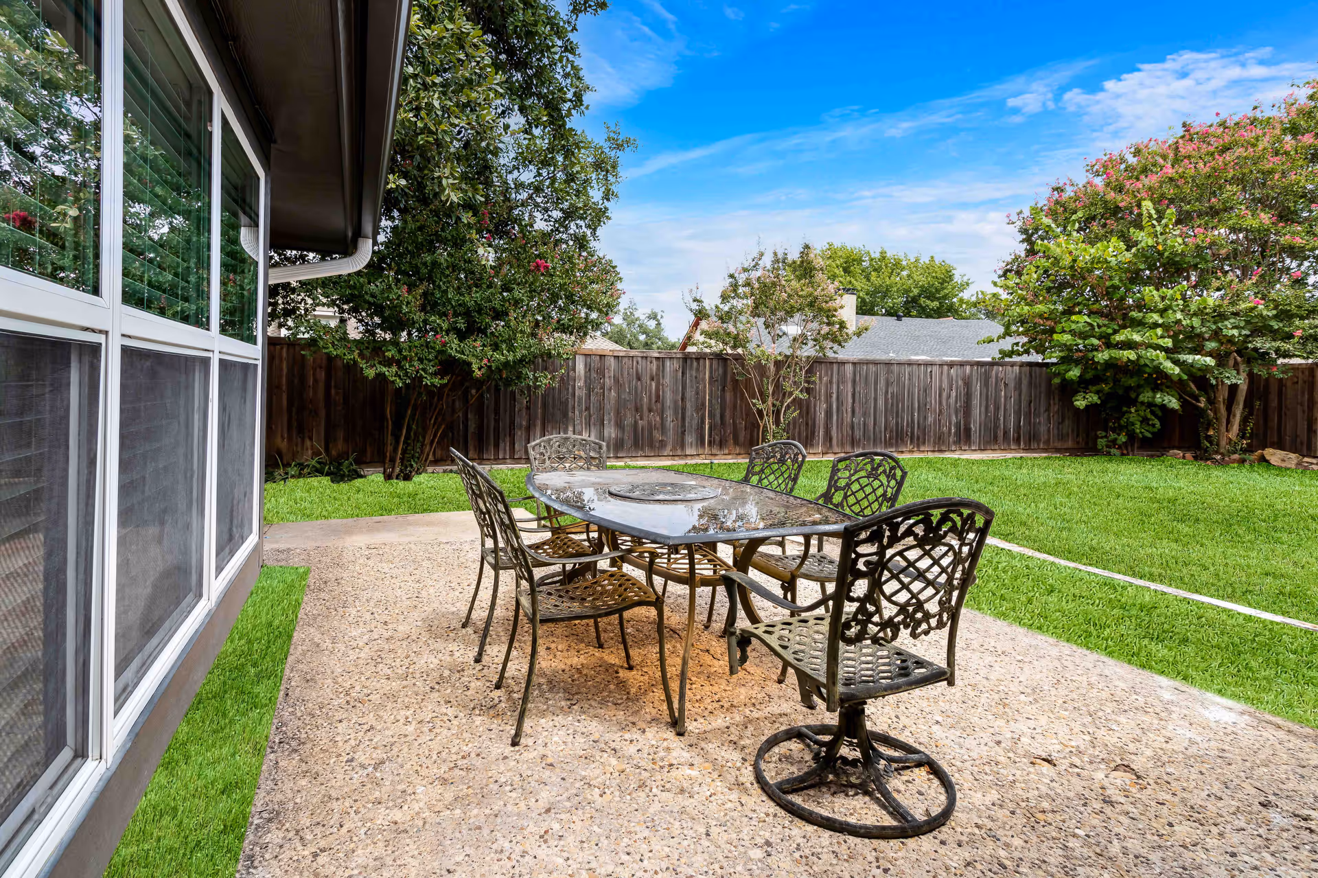 Outdoor patio area with a metal table and six matching chairs on a concrete surface, surrounded by green grass, trees, and a wooden fence under a blue sky with some clouds.