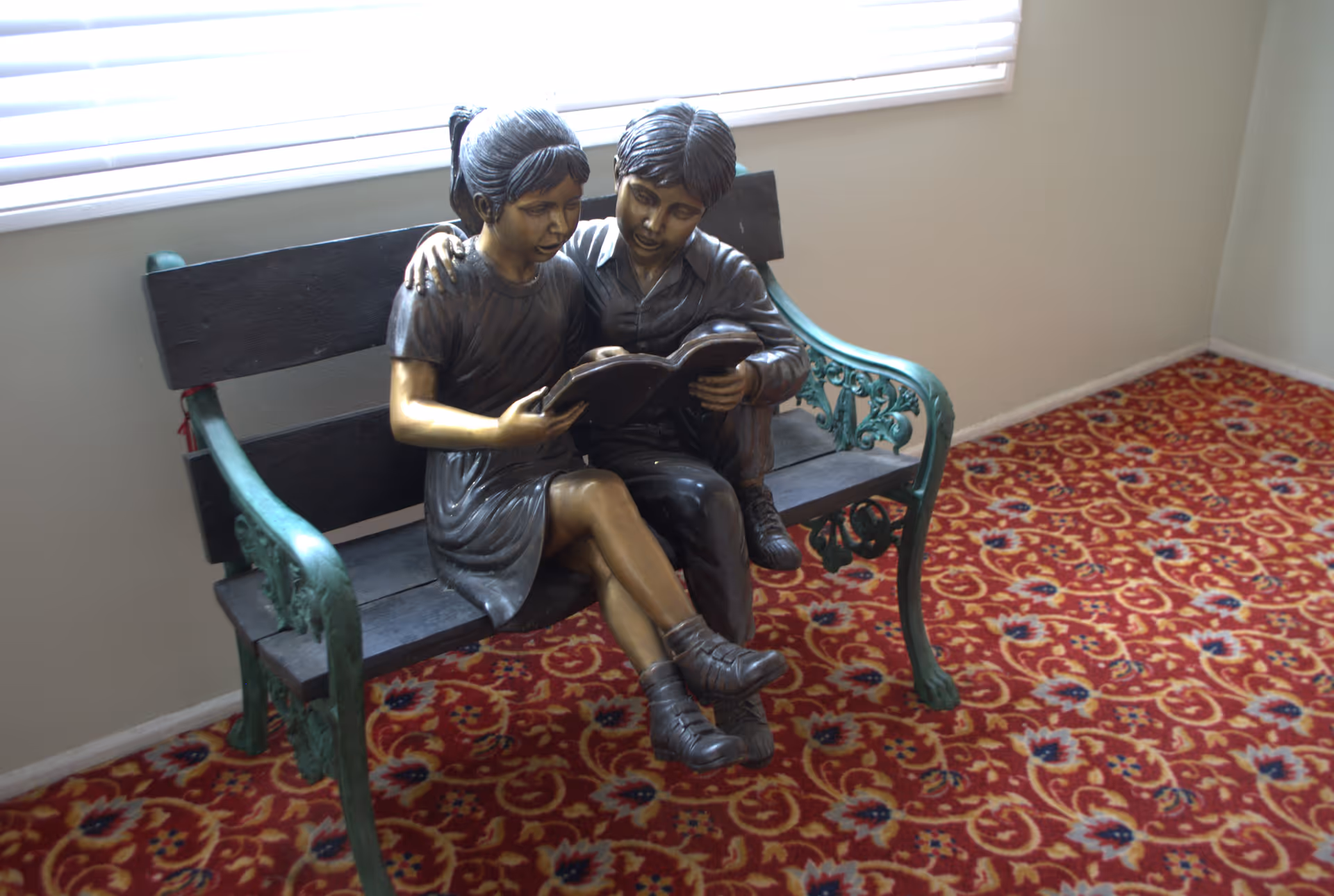 A bronze statue of two children sitting closely on a green metal bench with wooden slats, reading a book together. The bench is placed on a red patterned carpet in a room with beige walls and a window with white blinds.