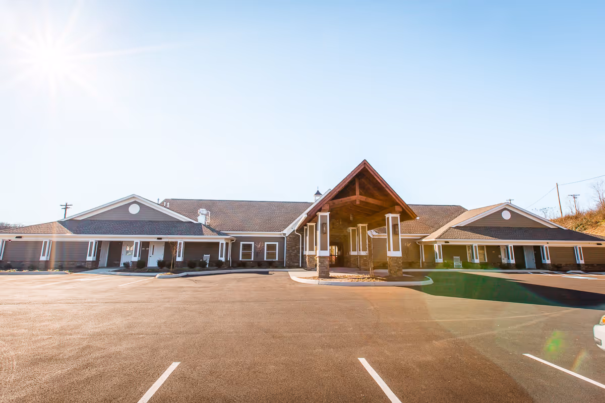 Exterior view of a single-story building with a large covered entrance and a spacious parking lot in front under a clear blue sky with the sun shining brightly.