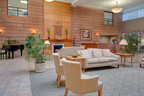 Spacious senior living lounge with sofas and chairs arranged around a coffee table, potted plants, a fireplace, grand piano and a reception desk.