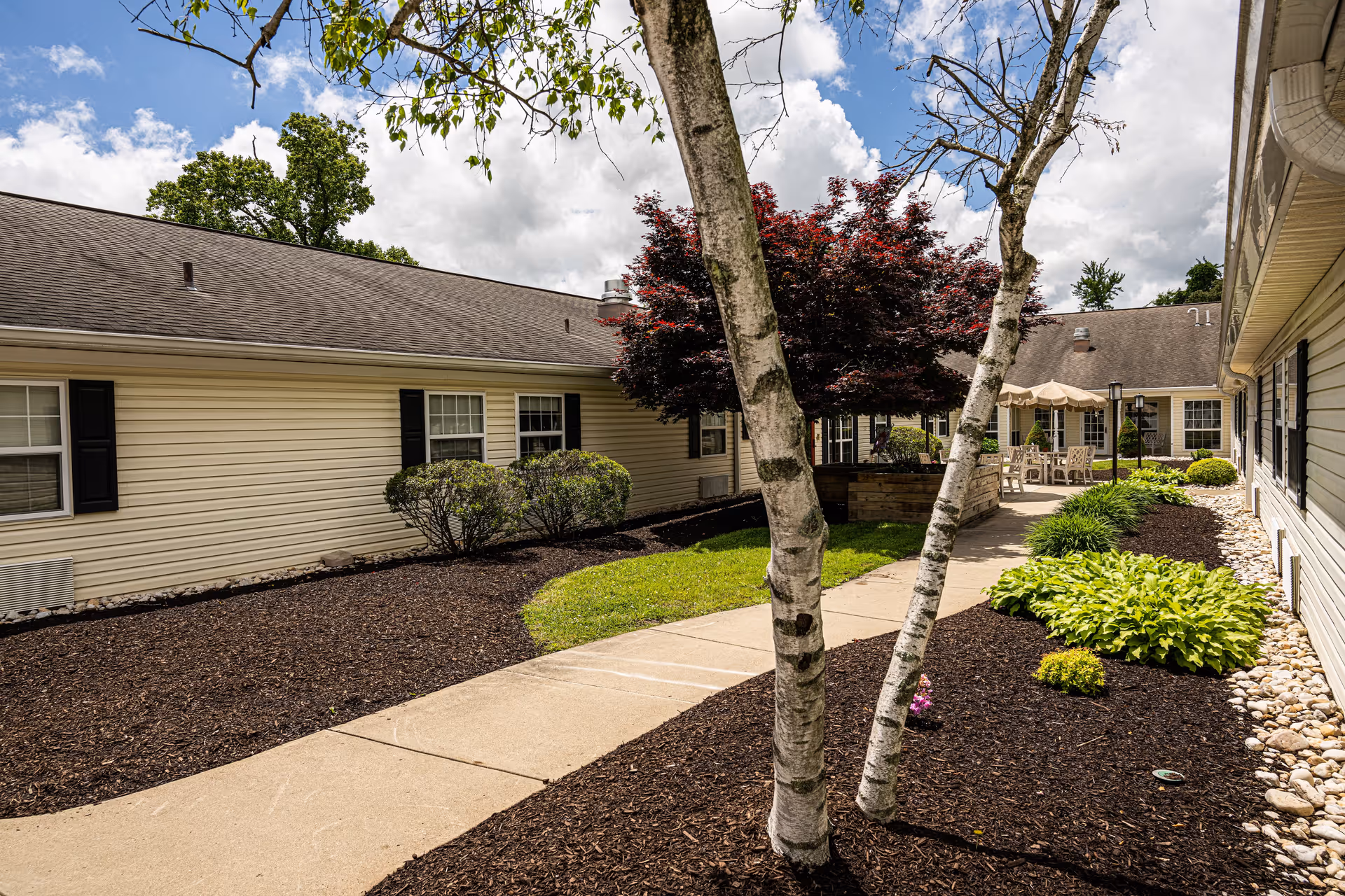Outdoor courtyard area of a senior living facility with a concrete walkway, landscaped garden beds with mulch, bushes, and trees. The building has beige siding with black shutters and multiple windows. There is a patio area with tables, chairs, and umbrellas in the background under a partly cloudy sky.