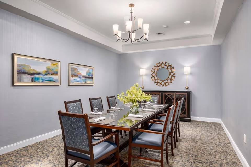 A formal dining room with a long rectangular glass-top table surrounded by eight wooden chairs with blue upholstered seats and backs. The table is set with white napkins, glasses, and a vase of yellow flowers in the center. The room has light gray walls, two framed landscape paintings on one wall, a decorative round mirror above a dark wooden sideboard, and two tall lamps on either side of the mirror. A chandelier with six lights hangs from the ceiling.