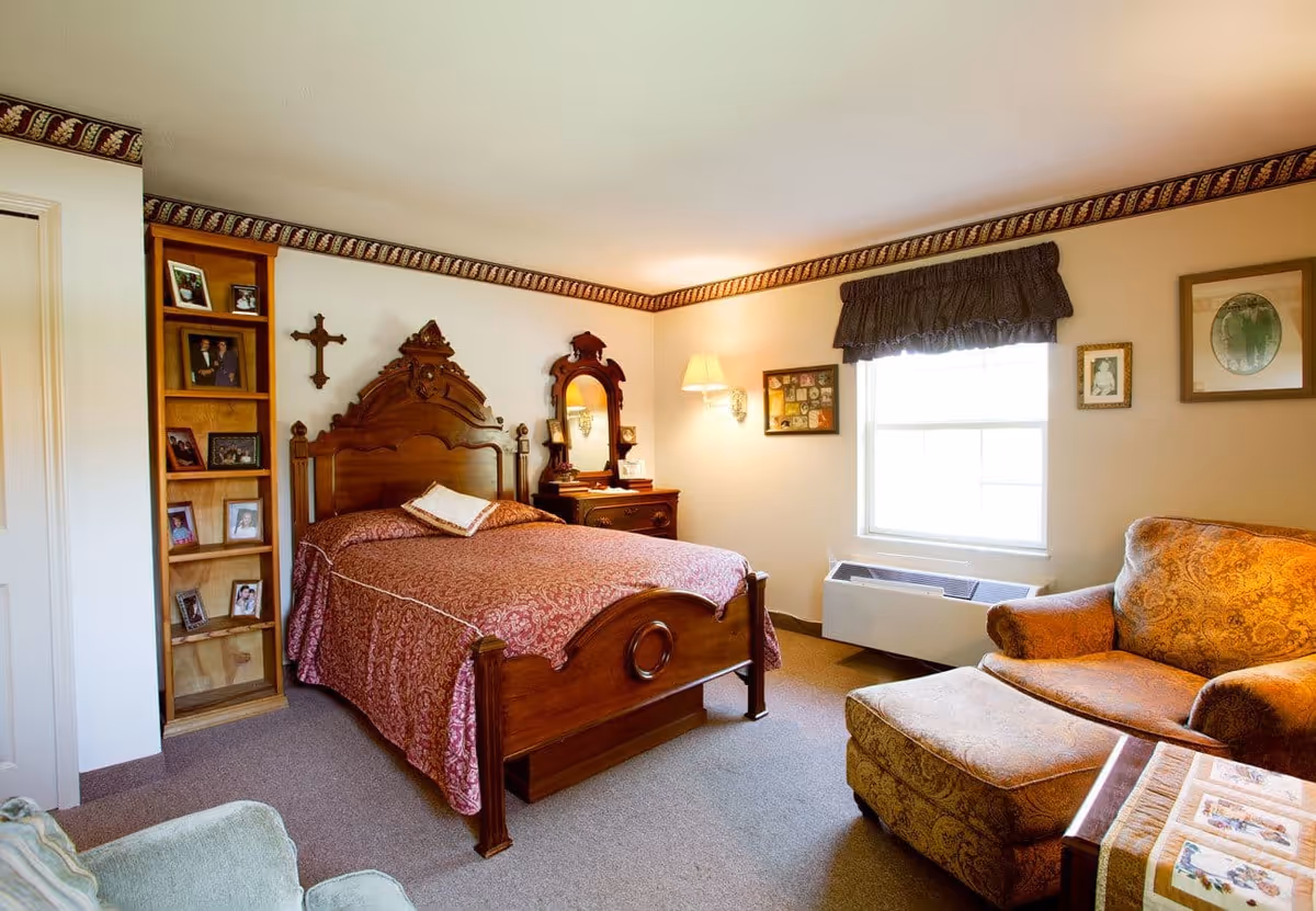 A cozy bedroom with a wooden bed frame and a red patterned bedspread. There is a wooden dresser with a mirror and a lamp on the wall above it. A window with a dark valance lets in natural light. The room also features a comfortable armchair with an ottoman and a small table. Family photos and framed artwork decorate the walls and a wooden shelf with more photos is next to the bed.