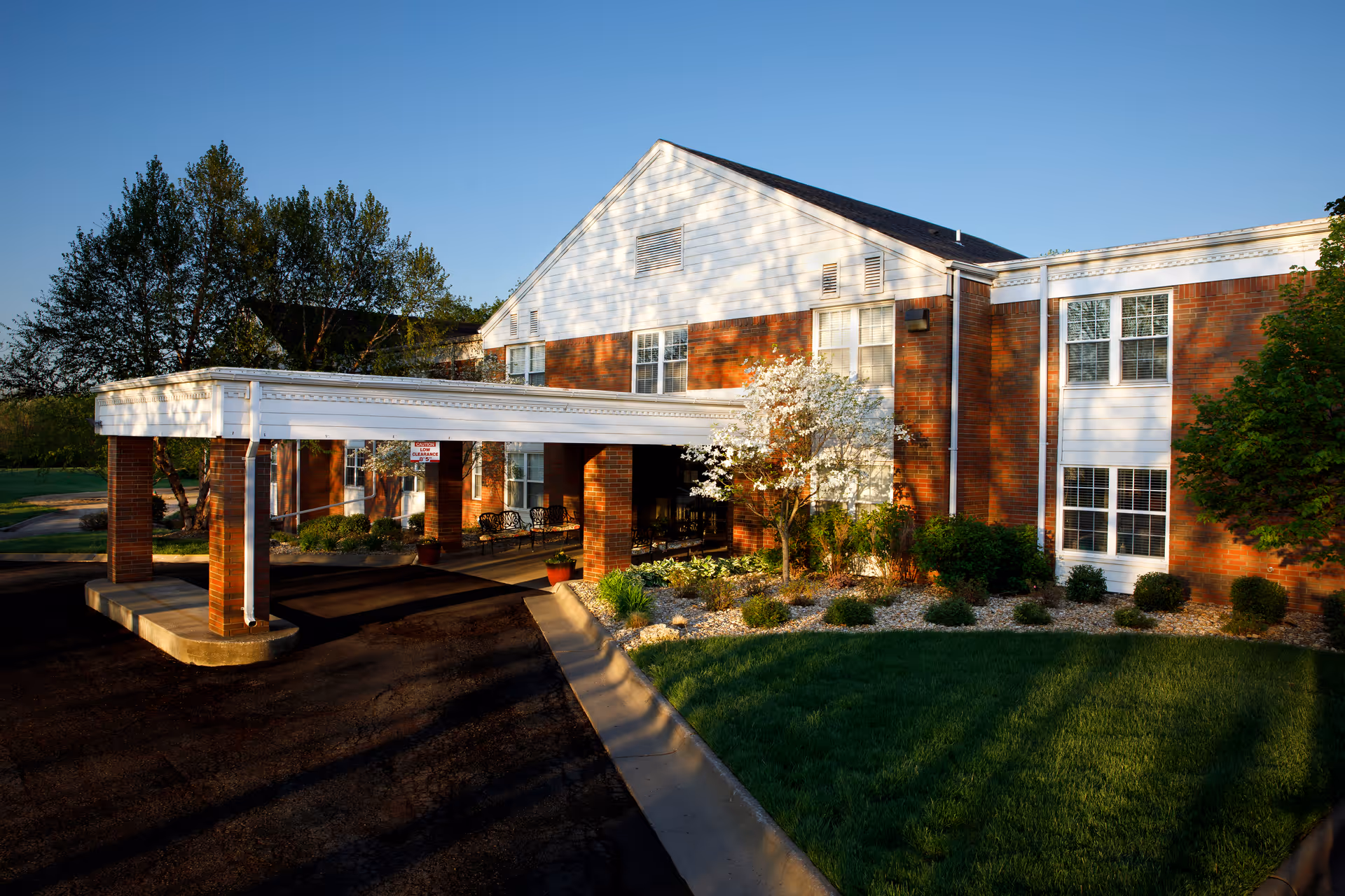 Entrance canopy and landscaped front of a two-story brick senior living building under a clear blue sky.