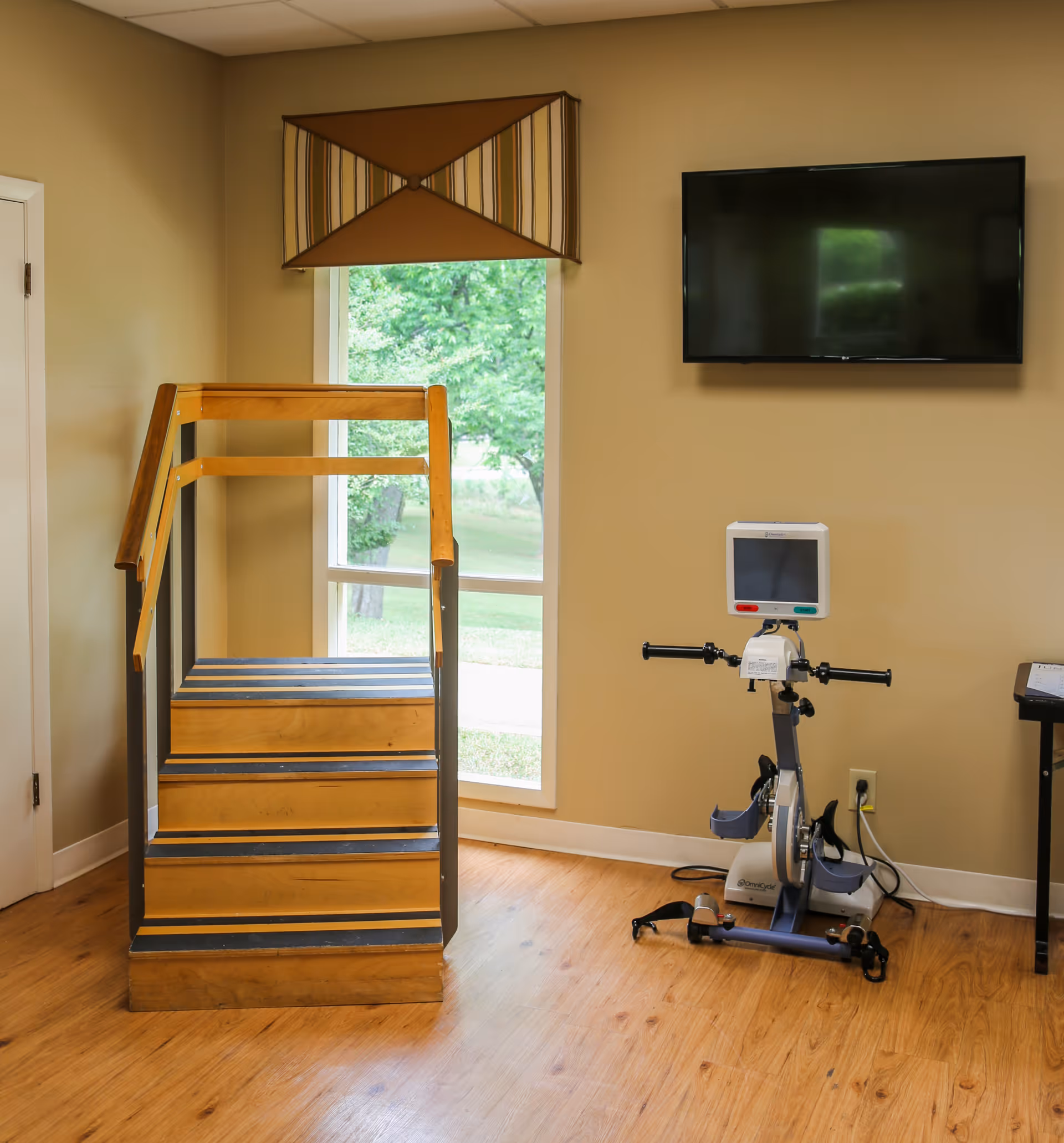 Small rehabilitation room with wooden practice stairs, a stationary exercise bike, and a wall-mounted TV beside a window.