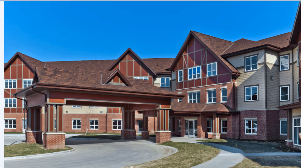 Exterior view of AVIVA Woodlands senior living facility showing a large multi-story building with a covered entrance driveway, red brick and beige siding, multiple windows, and a clear blue sky.