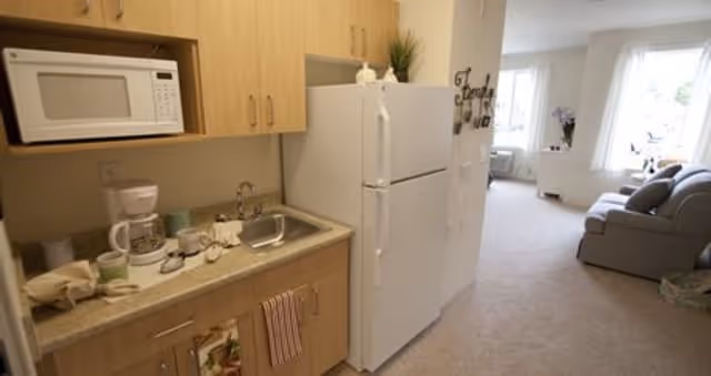 A small kitchen area with light wood cabinets, a microwave, a coffee maker, and a sink on the left side. A white refrigerator stands next to the kitchen, and beyond it is a carpeted living room with a gray sofa and a window letting in natural light.