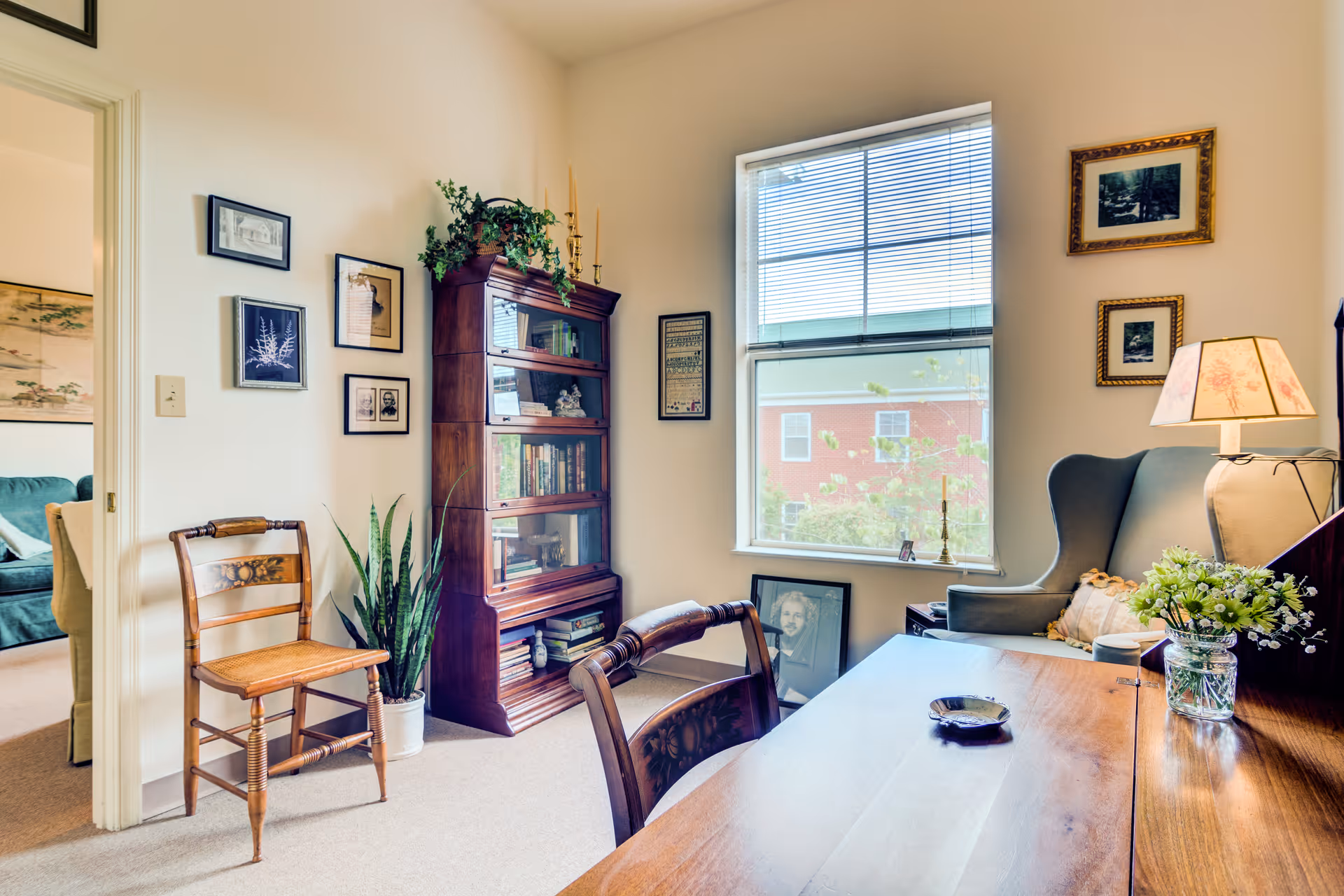 A cozy interior room with a wooden desk and chair in the foreground, a cushioned armchair with a floral lamp on a side table, a tall wooden bookshelf filled with books and decorative items, framed pictures on the walls, a potted plant on the floor, and a window with blinds letting in natural light.