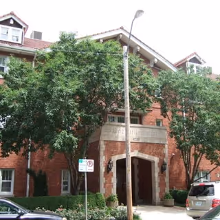 Exterior view of Rockhill Manor, a multi-story brick building with a covered entrance and several windows. Trees and shrubs are planted in front of the building, and a street lamp and parked cars are visible nearby.
