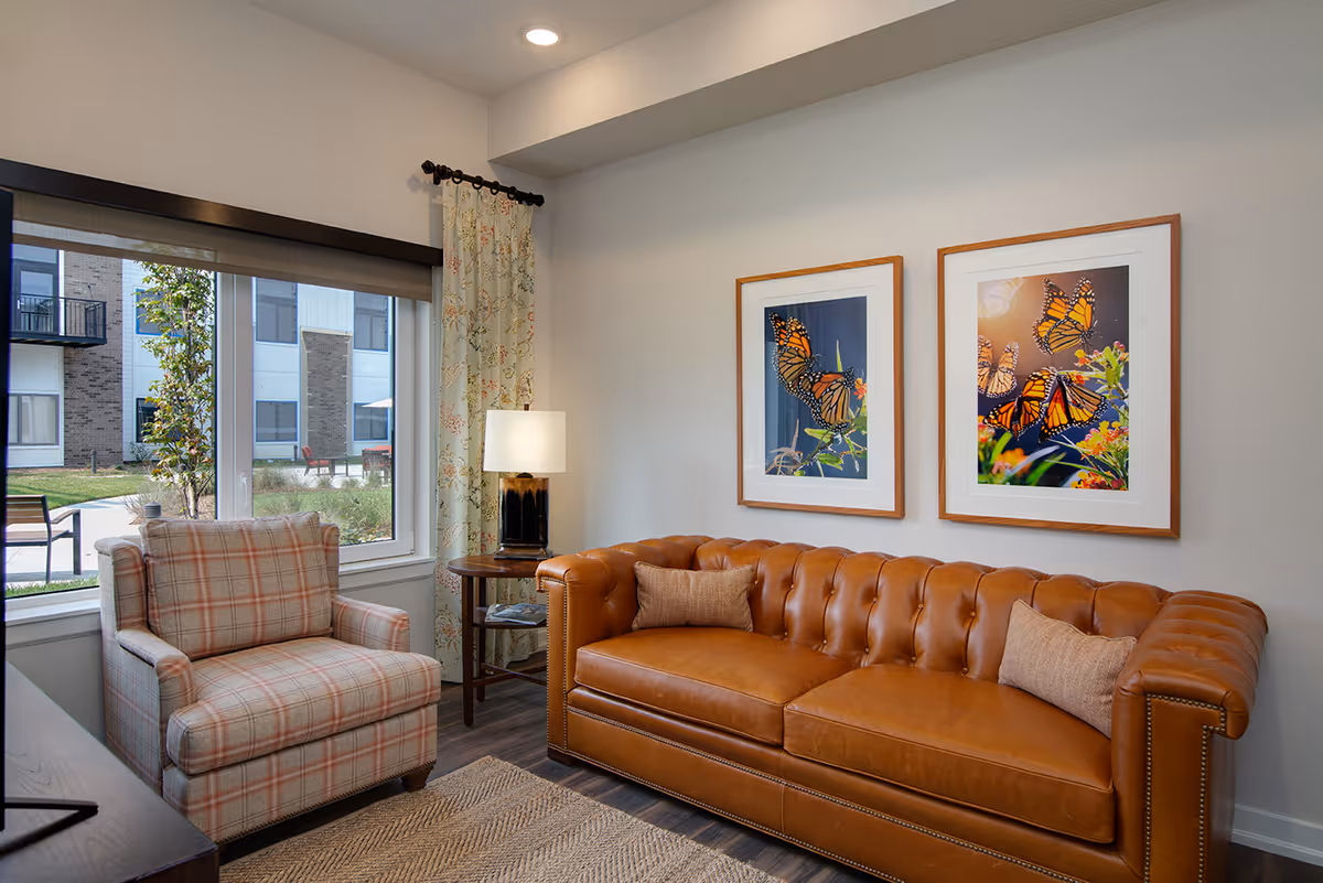 A cozy living room area with a brown leather tufted sofa adorned with two beige pillows, a plaid upholstered armchair, a wooden side table with a lamp, and two framed pictures of monarch butterflies on the wall. A large window shows an outdoor courtyard with greenery and building exteriors.