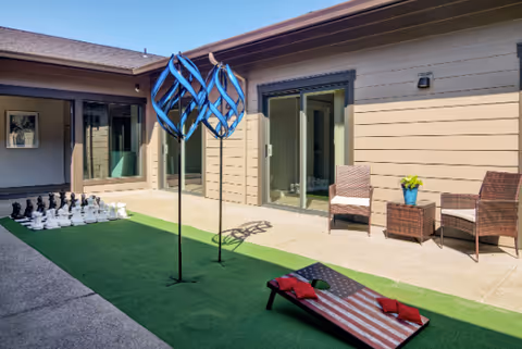 Outdoor courtyard area with artificial grass featuring a large chess set, two blue metal sculptures, a cornhole game with an American flag design, and two wicker chairs with a small table holding a potted plant. The area is surrounded by beige building walls with sliding glass doors.