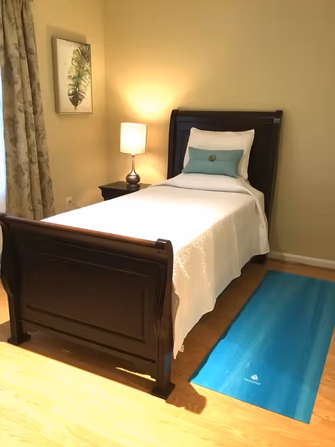A neatly made single bed with a dark wooden frame, white bedding, and a decorative pillow. Next to the bed is a nightstand with a lit lamp. A framed leaf artwork hangs on the wall above the nightstand. A blue yoga mat is placed on the wooden floor beside the bed. Beige curtains cover a window on the left side of the room.
