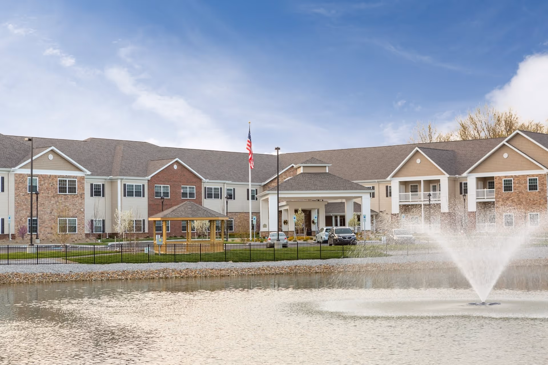 Exterior view of Danbury North Ridgeville senior living facility with a large pond and water fountain in the foreground, a gazebo, an American flag, and a two-story building with brick and siding under a partly cloudy sky.