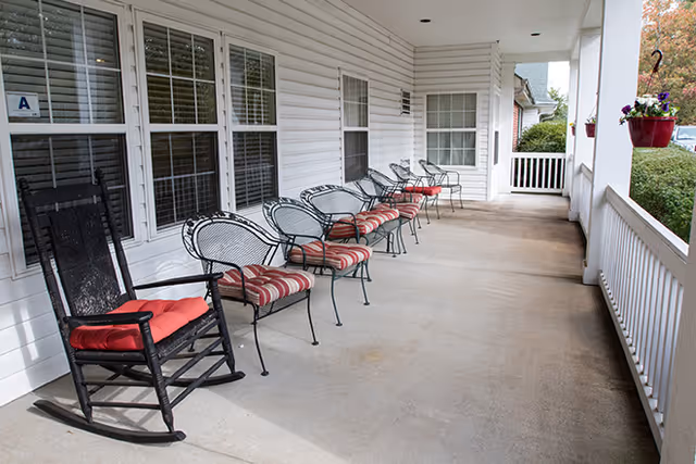 A covered outdoor porch area with a row of metal chairs with red and white striped cushions and a black rocking chair with a red cushion. The porch has white railings and hanging flower pots with purple flowers. The building exterior is white with multiple windows.