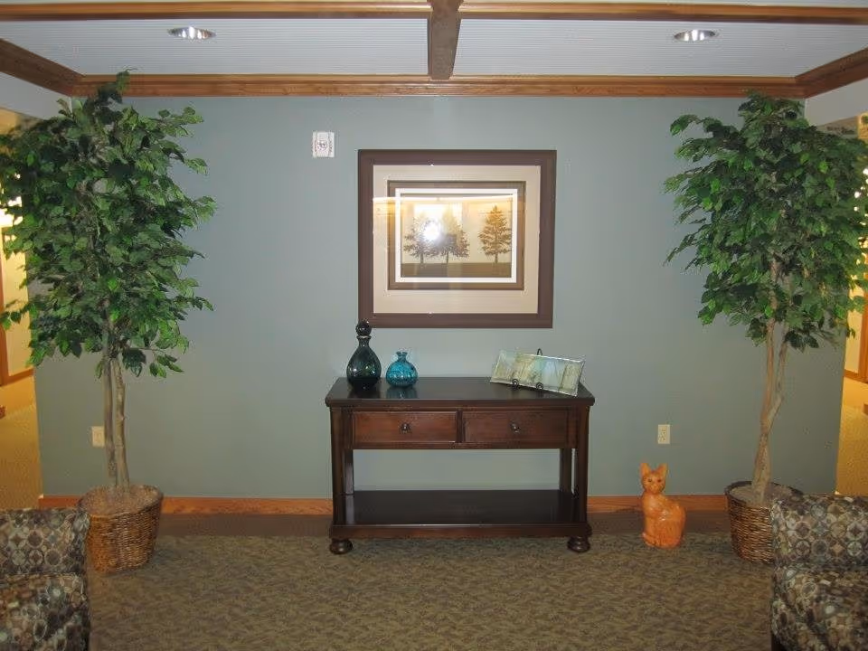 Interior view of a hallway or common area in Hansen House featuring a wooden console table with two drawers. On the table are decorative glass bottles and a framed item. Above the table hangs a framed picture of trees. Two large potted plants flank the table on either side. A small orange cat figurine is placed on the floor to the right of the table. The walls are painted green with wood trim, and the ceiling has recessed lighting.