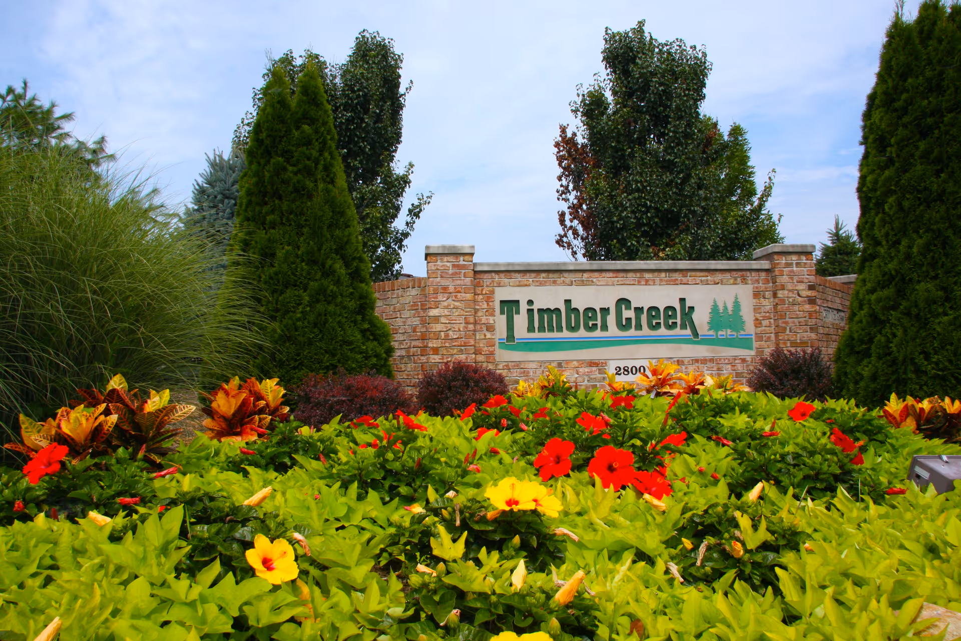 A landscaped garden area with colorful flowers and green shrubs in front of a brick wall sign that reads 'Timber Creek' with a small tree logo and the number 2800 below it. Tall trees are visible behind the wall under a partly cloudy sky.
