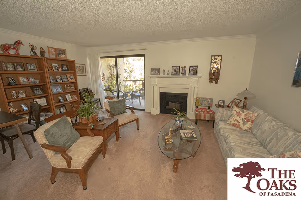 A cozy living room with a beige carpet, a patterned sofa with floral cushions, two wooden armchairs with green cushions, a glass coffee table with decorative items, a white fireplace with framed photos on the mantel, a wooden bookshelf filled with framed pictures, and a sliding glass door leading to a balcony with outdoor chairs and plants.