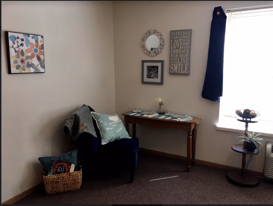 A cozy corner of a room with a dark blue armchair draped with a patterned throw and a light blue pillow. Next to the chair is a woven basket containing books and another pillow. A wooden console table against the wall holds a small vase with a flower and some decorative items. Above the table are three wall decorations: a round ornate mirror, a framed picture, and a sign with inspirational words. To the right, a window with a dark curtain lets in natural light, and a small round wooden stand holds a plant and a decorative bowl.