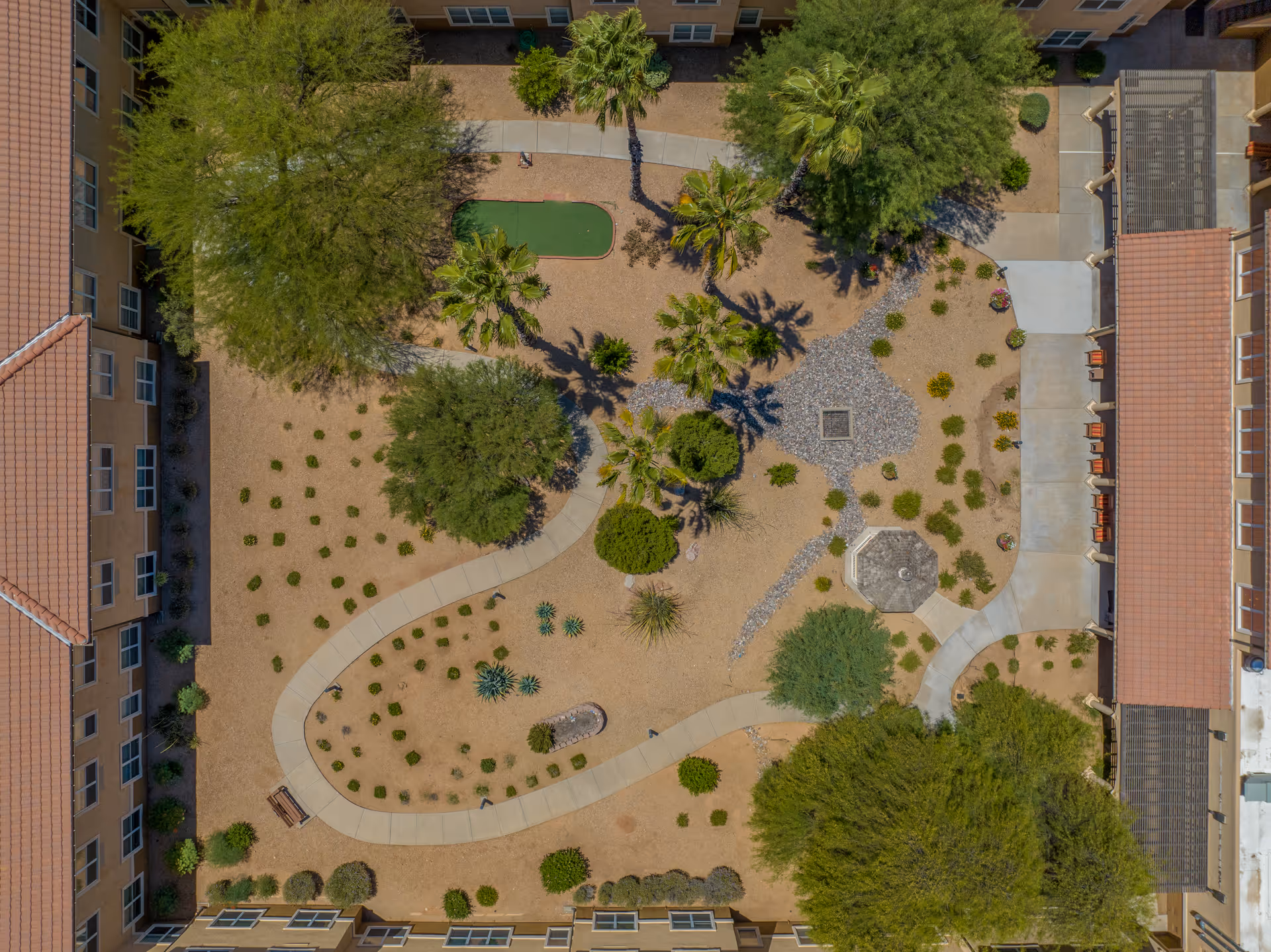 Aerial view of a landscaped courtyard at Sierra Del Sol Assisted Living, featuring walking paths, trees, shrubs, a small putting green, and a gazebo surrounded by a building with tiled roofs.